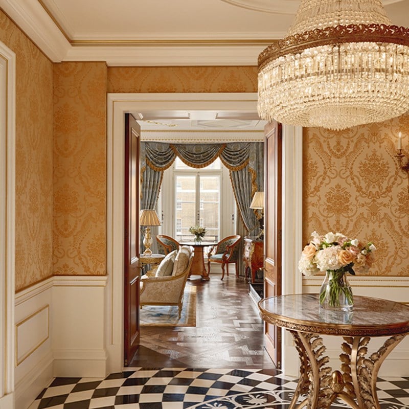 Entrance hall with crystal chandelier, floral arrangement on round table, and view through double doors to a richly decorated sitting room.
