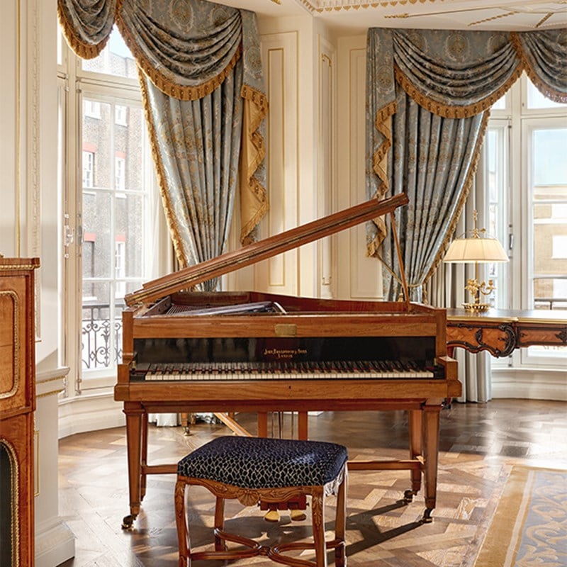 Antique grand piano and velvet stool beside tall windows draped in blue and gold curtains, set on polished parquet floors.