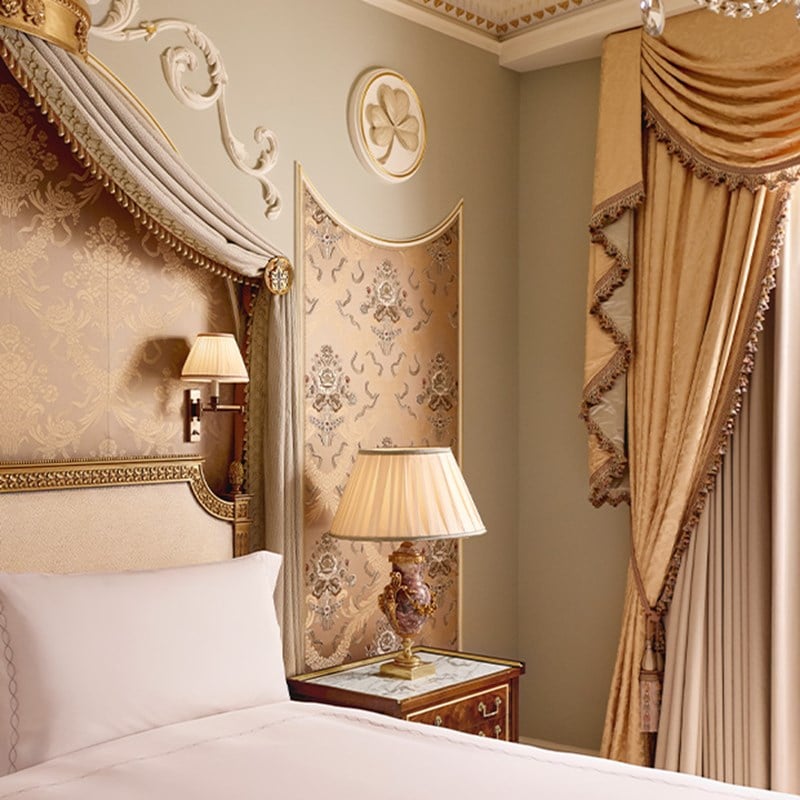 Bedroom corner with gold-accented headboard, embroidered panels, classical wall mouldings, and warm lighting from a marble and bronze bedside lamp.