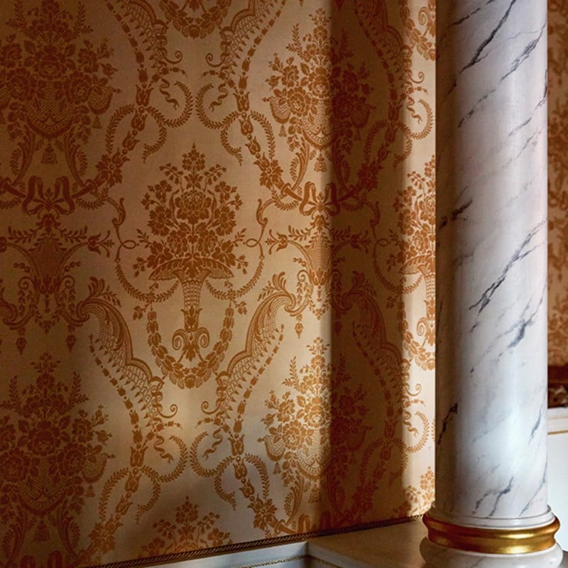 Close-up of traditional damask wallpaper in golden tones, softly lit, beside a white faux-marble pillar with gold base detailing.