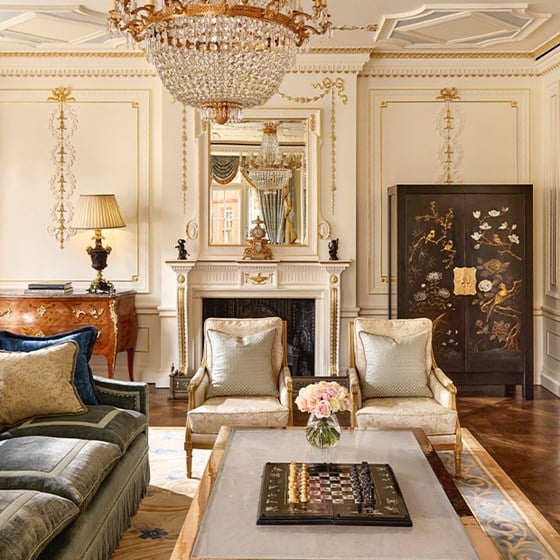 Elegant salon with chandelier, marble fireplace, gilded mirror, pale armchairs, and coffee table topped with fresh flowers and a chess set.
