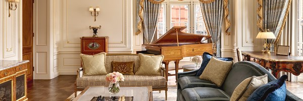 Luxurious lounge with grand piano, blue and gold curtains, carved wooden writing desk, and silk-upholstered seating in soft neutral tones.