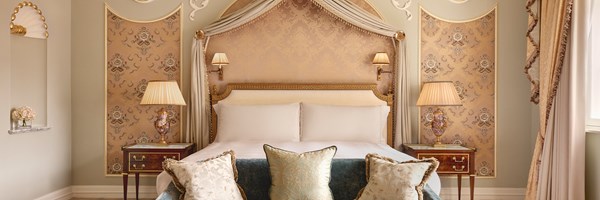 Regal bedroom with canopied headboard, gold-trimmed nightstands, crystal lamps, and velvet furnishings beneath ornate cornicing and pleated drapes.