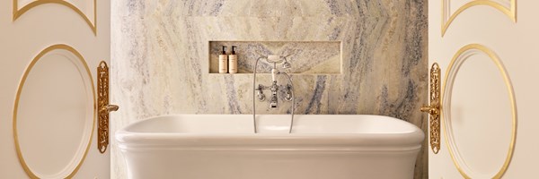 Freestanding bathtub framed by double doors with gold handles, set against softly veined marble wall and recessed shelf with toiletries.