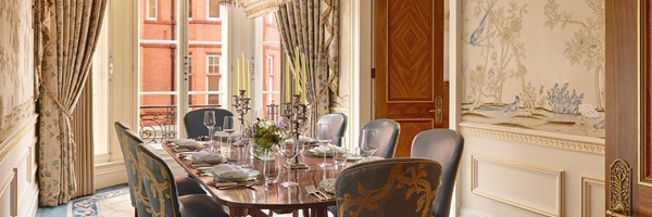 Elegant dining room with polished oval table, blue and gold chairs, floral centrepiece, silver candelabras, and hand-painted wallpaper with birds.