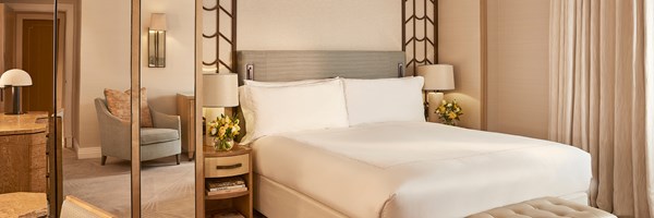 Bedside detail of Superior Room showing a velvet-upholstered headboard, wood and leather side table with a lamp, and a small floral arrangement beside the crisp white bed linen.