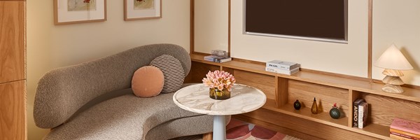 A cosy lounge corner with a curved grey sofa, round marble-topped table with pink flowers, and built-in wooden shelving decorated with books, vases, and a sculptural lamp beneath a wall-mounted TV.