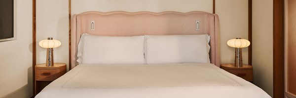 A soft-toned bedroom featuring a pink upholstered headboard, white bed linens, and rounded bedside tables topped with sculptural lamps, framed by vertical wooden panels.