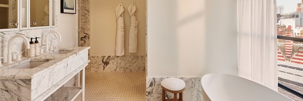 A light-filled marble bathroom with dual sinks, luxury toiletries, two hanging robes, a freestanding bathtub, and a window offering views of London rooftops.
