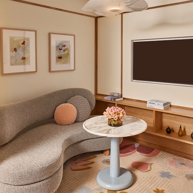 A cosy lounge corner with a curved grey sofa, round marble-topped table with pink flowers, and built-in wooden shelving decorated with books, vases, and a sculptural lamp beneath a wall-mounted TV.