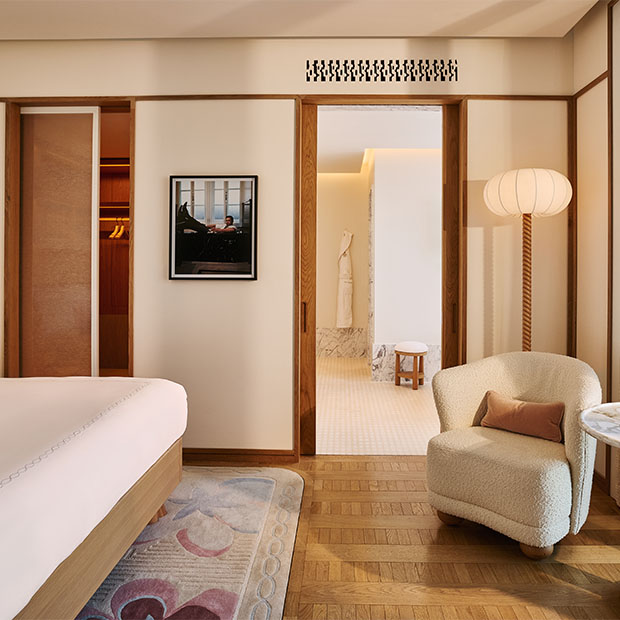 A view from the bedroom into the bathroom showing a boucle armchair, a sliding wooden door, a hanging robe, and tiled flooring leading to a freestanding tub.