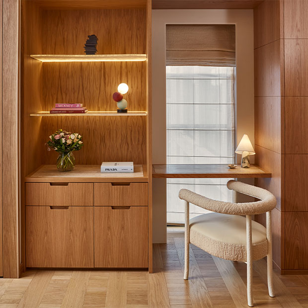 A built-in wooden desk area with a modern upholstered chair, open shelving displaying books, flowers, and decorative lighting.