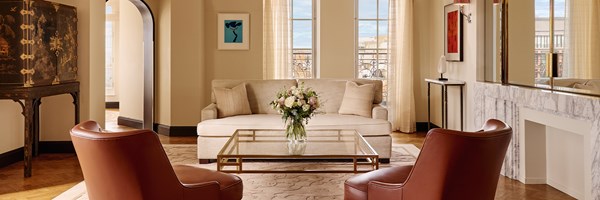 Elegant living room with cream sofa, tan leather armchairs, glass coffee table, and tall French doors opening onto a balcony with skyline views.