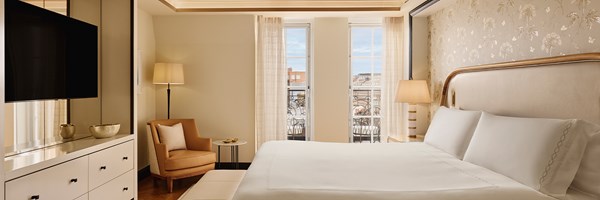 Refined bedroom with king-size bed, floral feature wall, soft cream tones, and French doors revealing city rooftops.