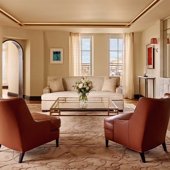 Symmetrical living room with two tan leather armchairs facing a cream sofa, glass table, floral arrangement, and marble fireplace.