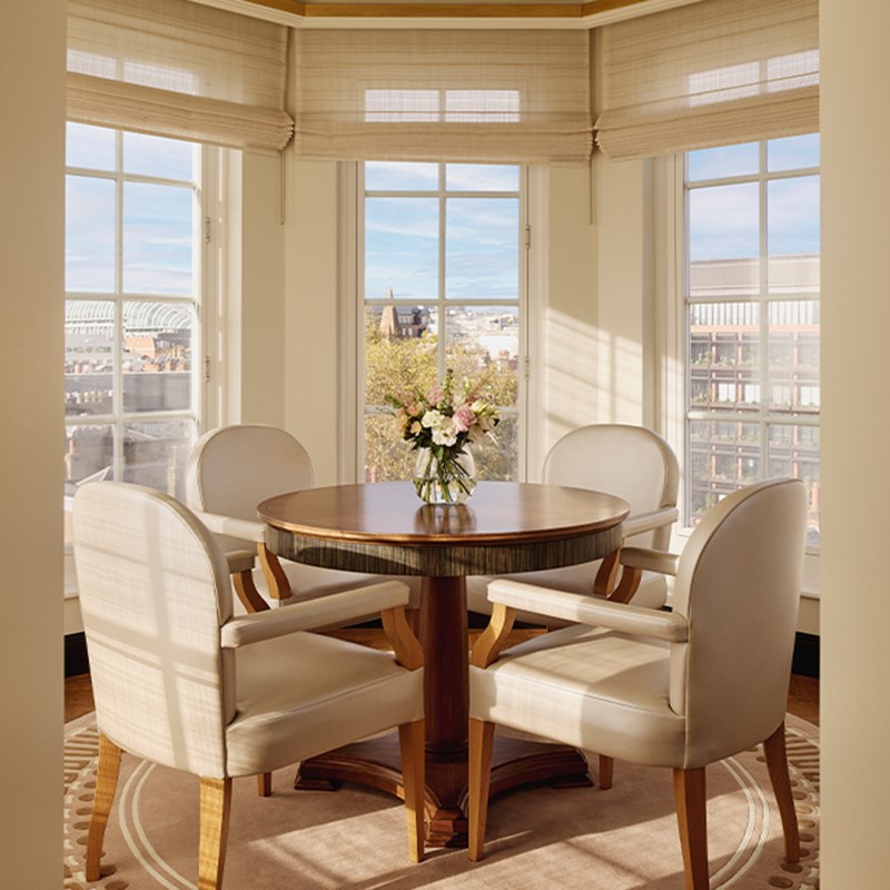 Intimate dining space with round wooden table and four upholstered armchairs surrounded by panoramic city views through curved windows.