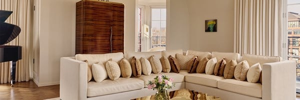 Bright living room with cream L-shaped sofa, gold and bronze cushions, floral arrangement, and large windows opening onto balcony.