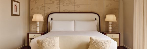 Calming bedroom with cream upholstered bed, twin gold-striped lamps, and illustrated wall panels featuring birds and clouds in warm tones.