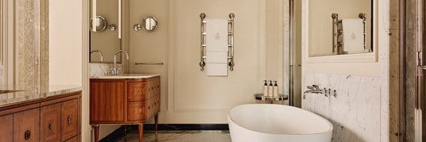 Classic marble bathroom with freestanding tub, wooden vanity units, chrome fixtures, and luxury toiletries neatly arranged on a chrome stand.