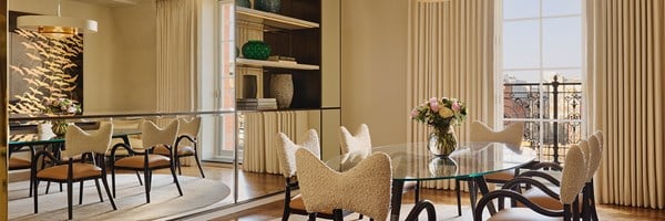 Light-filled dining area with city views, sculptural chairs, glass table, mural wall, and decorative shelving featuring vases and art books.