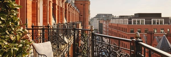 Ornate black wrought iron balcony with leafy planting, cream lounge chair, and views of red brick façades and central London rooftops.