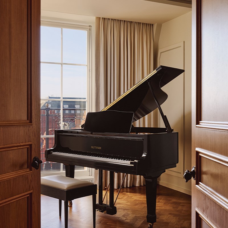Elegant music room with black grand piano and stool, framed by full-height windows and soft beige curtains.