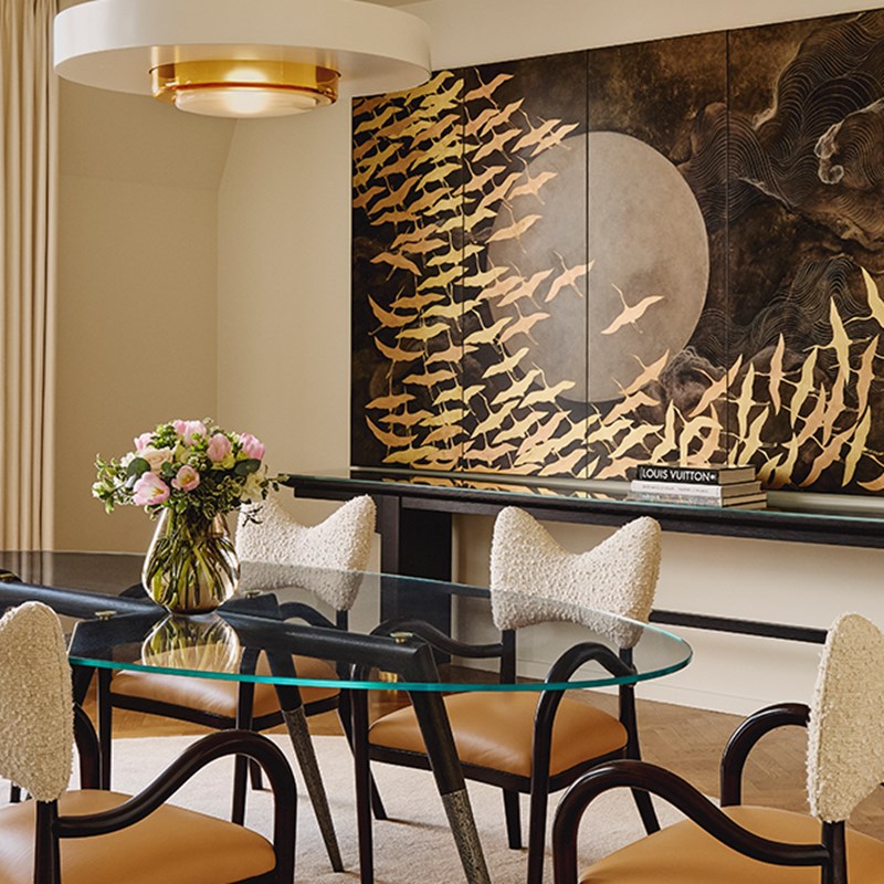 Contemporary dining area with glass table, boucle chairs, floral centrepiece, and gold bird mural set against a moonlit backdrop.