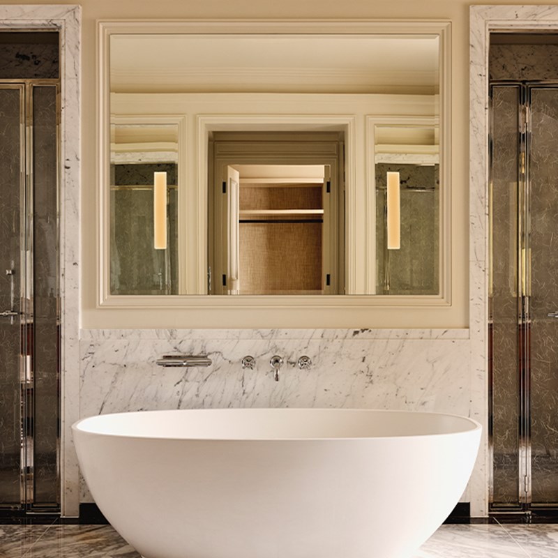 Freestanding oval bathtub set against a marble wall, facing a large mirror with soft lighting and double walk-in showers on either side.