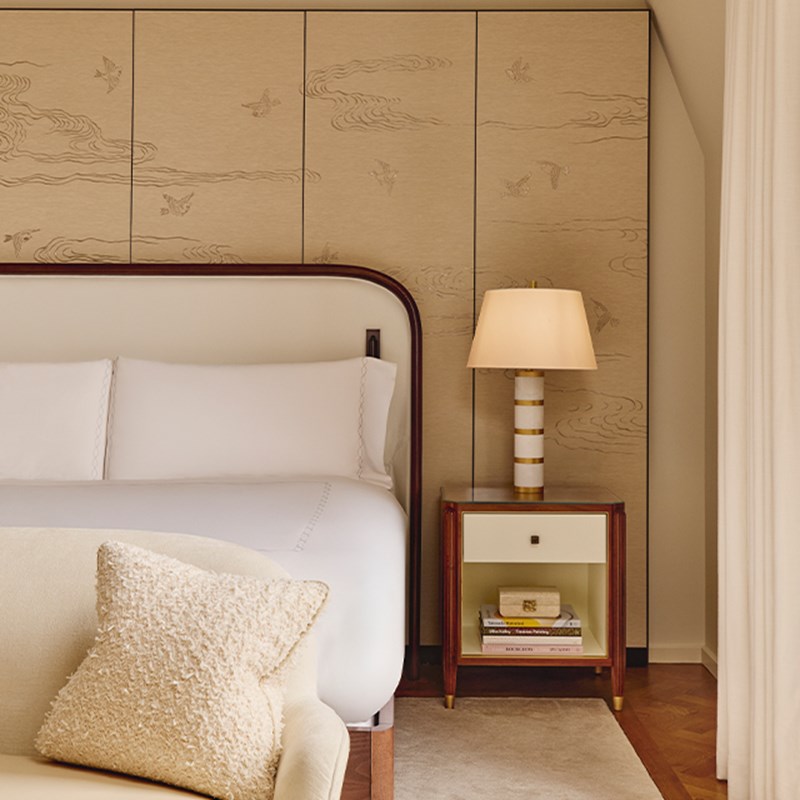 Close-up of cream-upholstered bed with curved headboard, striped lamp on wooden side table, and wall panels etched with birds and clouds.