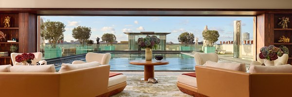 Luxurious lounge area opens onto rooftop terrace with swimming pool, sculpted trees, and skyline views under a bright blue sky.
