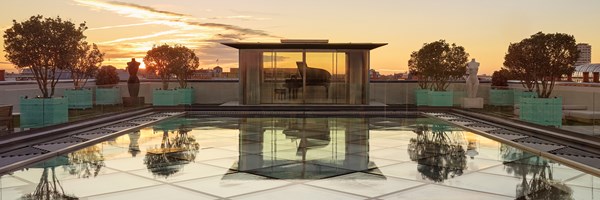 Rooftop terrace at sunset with glass pool, sculpture-lined edges, and a grand piano enclosed in a modern glass pavilion.