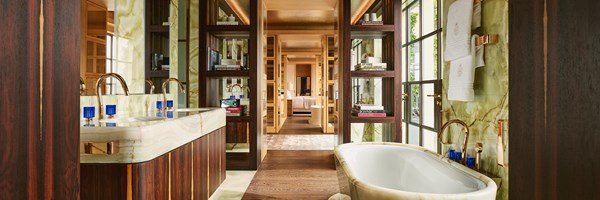 Luxurious bathroom with twin marble sinks, freestanding bathtub, bronze fixtures, and backlit shelving, looking through to a serene bedroom.