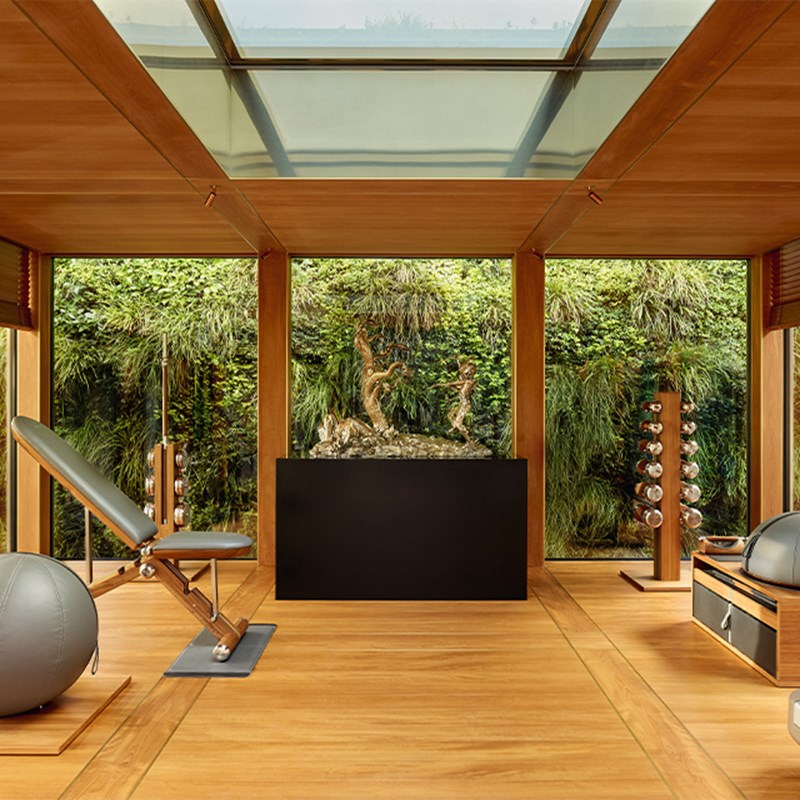 Elegant private gym with wood flooring, skylight ceiling, mirrored equipment, and lush green vertical garden wall beyond floor-to-ceiling windows.