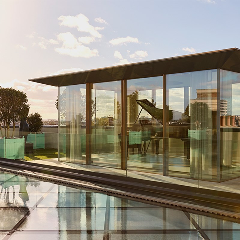 Modern rooftop glass pavilion with grand piano inside, reflecting on water feature beneath a bright sky with scattered clouds.