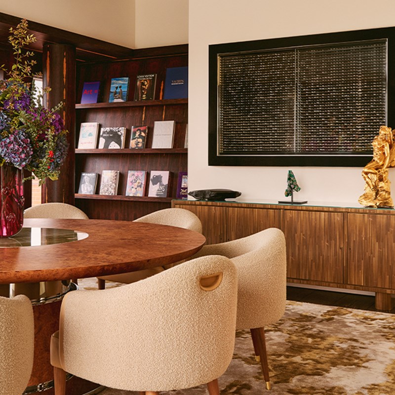 Stylish library or study with round wooden table, plush chairs, art books on dark shelves, and decorative sculptures on sideboard.