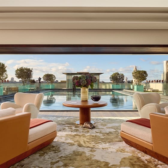 Luxurious lounge area opens onto rooftop terrace with swimming pool, sculpted trees, and skyline views under a bright blue sky.
