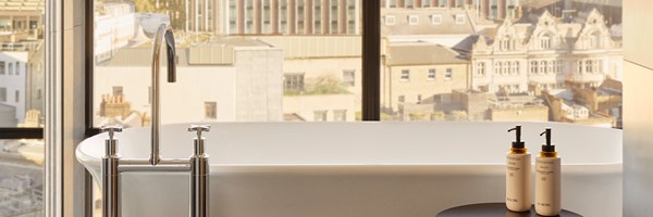 Modern freestanding bathtub beside full-height windows with skyline view, flanked by luxury Surenne bath products on a sleek side table.