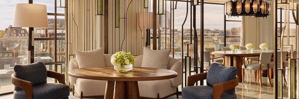 Stylish open-plan seating area with round wooden table, soft armchairs, decorative glass dividers, and expansive views across city rooftops.