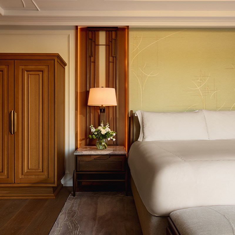 Calming bedroom corner with crisp white bed, soft green textured wall, floral arrangement, and symmetrical wooden cabinetry and sconce lighting.