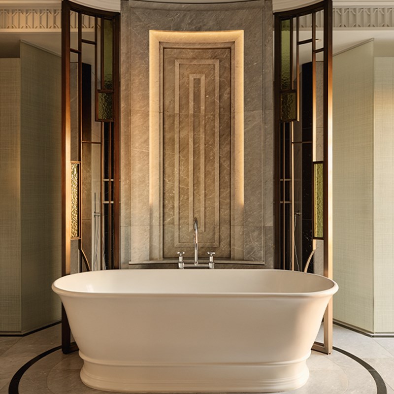 Freestanding white bathtub framed by marble panels and art deco-inspired lighting, with ambient lighting inset into a textured stone wall.