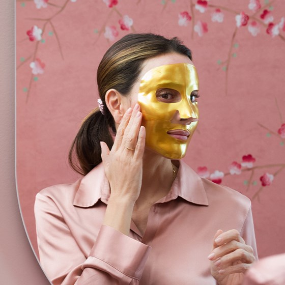 Woman looking at her reflection of herself in a gold MZ skin face mask. The background is pink.
