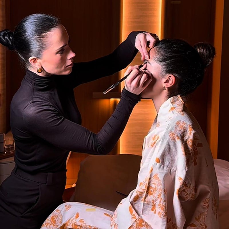 Makeup artist in a black turtleneck carefully applying eyeliner to a seated woman wearing a white robe with orange floral patterns, in a softly lit room with warm tones.