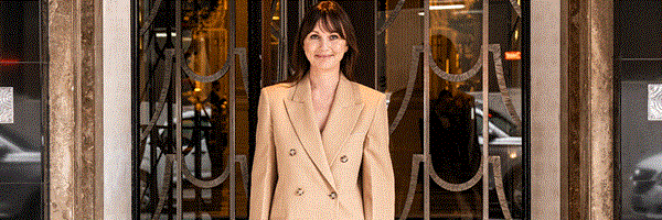 A woman in a beige double-breasted suit stands smiling at the entrance of Claridge’s, framed by art deco doors, with “CLARIDGE’S” visible on the doormat beneath her feet.