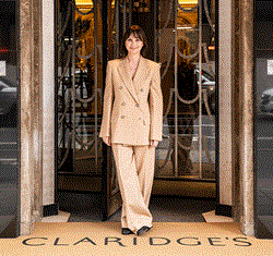 A woman in a beige double-breasted suit stands smiling at the entrance of Claridge’s, framed by art deco doors, with “CLARIDGE’S” visible on the doormat beneath her feet.