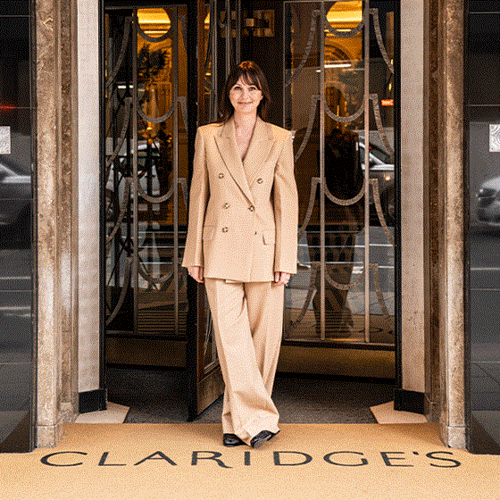 A woman in a beige double-breasted suit stands smiling at the entrance of Claridge’s, framed by art deco doors, with “CLARIDGE’S” visible on the doormat beneath her feet.
