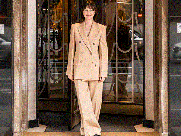 A woman in a beige double-breasted suit stands smiling at the entrance of Claridge’s, framed by art deco doors, with “CLARIDGE’S” visible on the doormat beneath her feet.