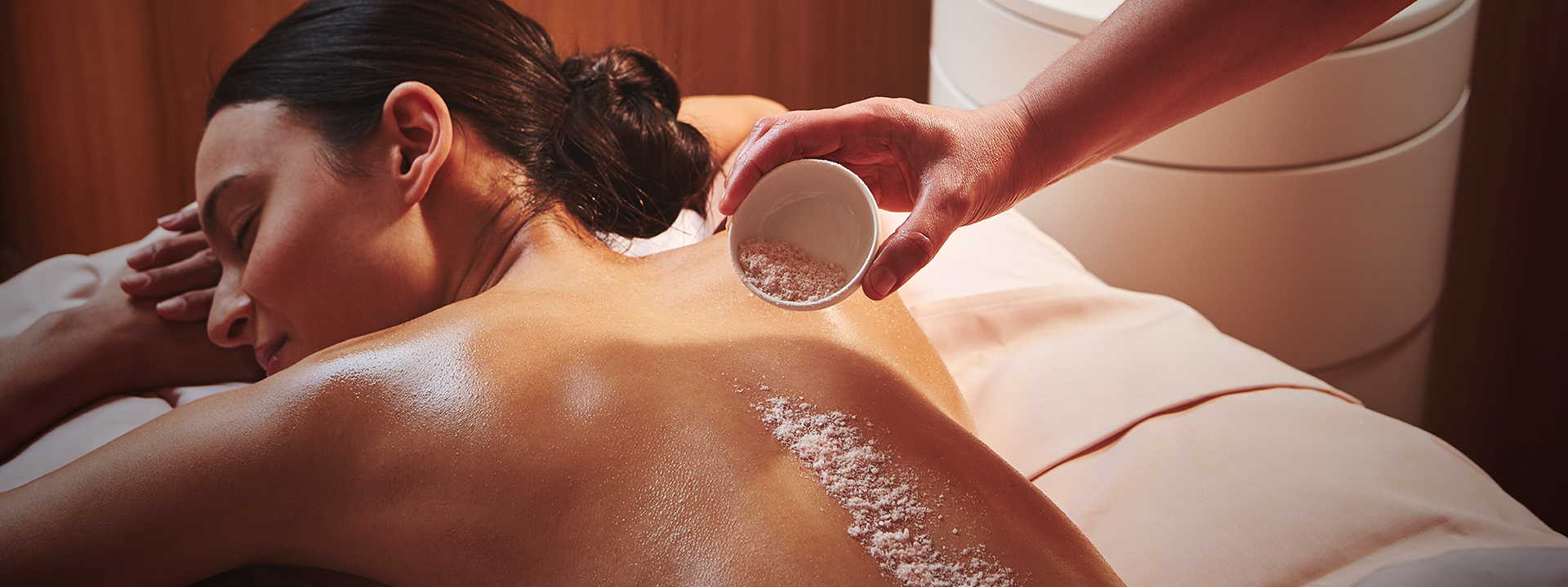 A girl lying in a relaxing La Eva body spa treatment and a hand applying body scrub to her back.