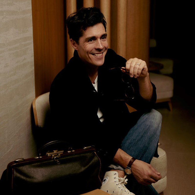 Ross smiling in a navy blue jacket, white t-shirt, blue jeans and white converses