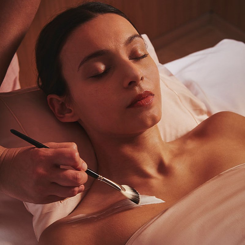 Woman lying with eyes closed as a therapist brushes a cream mask onto her upper chest.