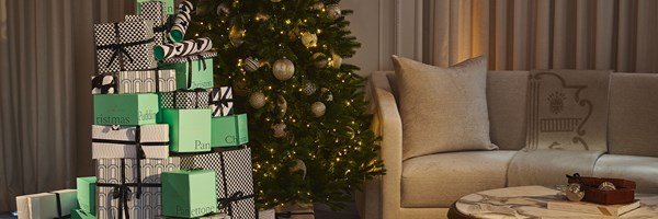 A stack of patterned and green Claridge’s Christmas gift boxes beside a decorated Christmas tree, with a beige sofa and marble coffee table in the background.